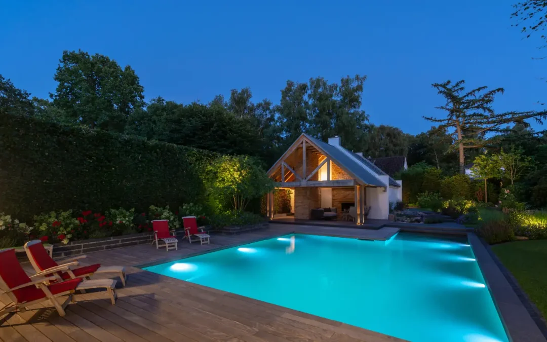 Im abendlichen Garten leuchtet der Pool neben dem Poolhaus, während rote Liegestühle, üppige Bepflanzung und warmes Licht Ruhe schaffen.