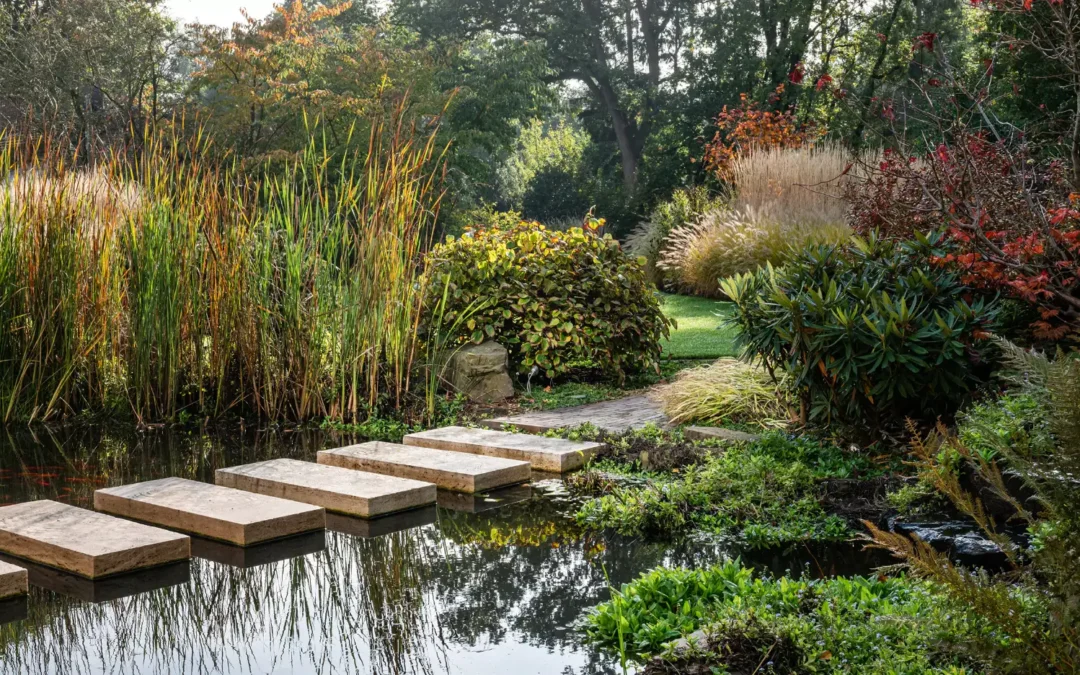 Im Garten von Beran Gärten führt ein geschwungener Weg mit Trittsteinen am Teich vorbei durch üppige Stauden, Gräser und Herbstfarben am Waldrand.