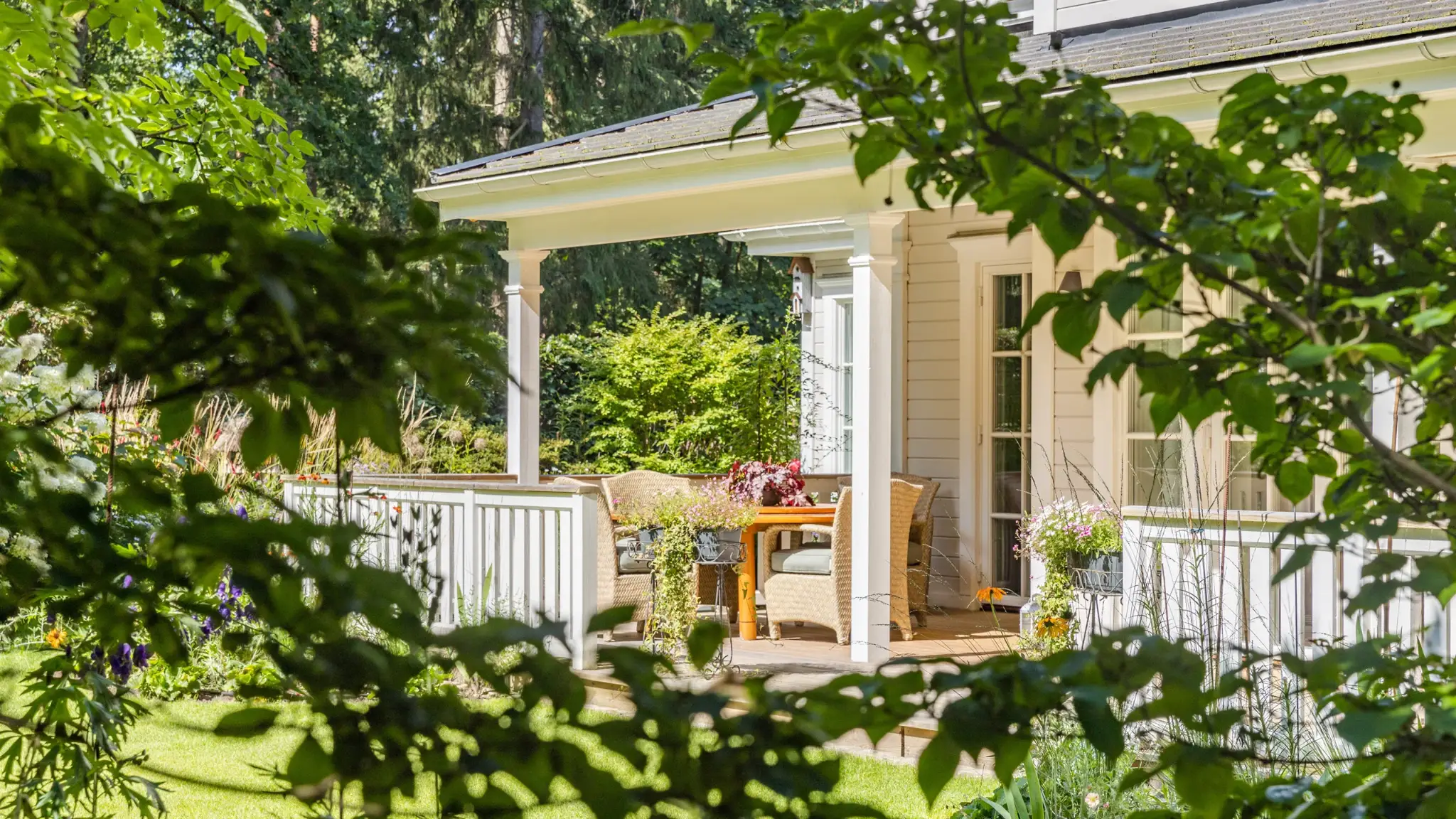 Durch dichtes Grün blickt man auf eine sonnige Hausveranda mit Rattanstühlen, Holztisch und bunten Blumen vor einem hellen Holzhaus im Garten.