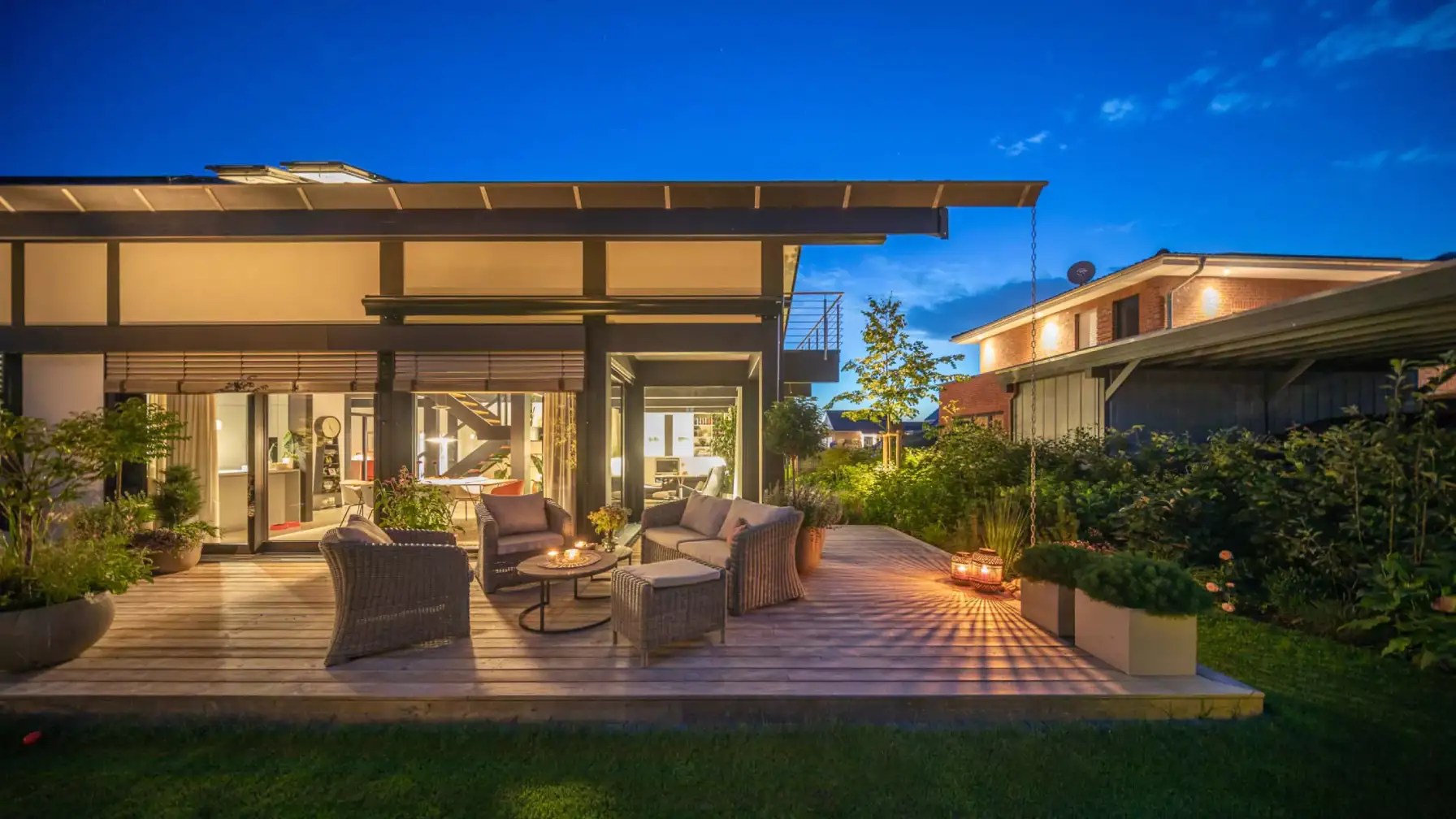 Auf der beleuchteten Holzterrasse am modernen Haus stehen Lounge-Möbel, Pflanzenkübel und Kerzenlicht unter dem tiefblauen Abendhimmel.