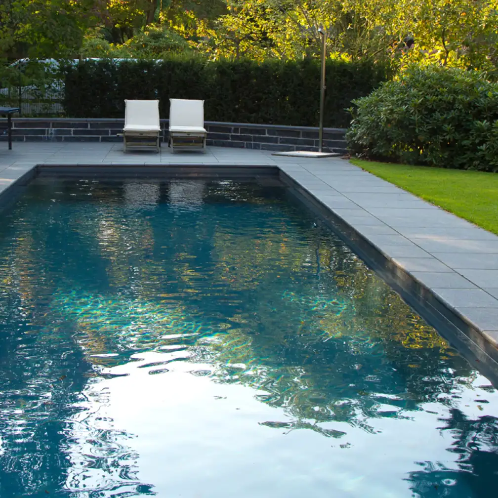 Im ruhigen Garten von Beran Gärten glänzt ein moderner Pool mit dunklem Rand neben zwei weißen Liegestühlen und gepflegter Rasenfläche.