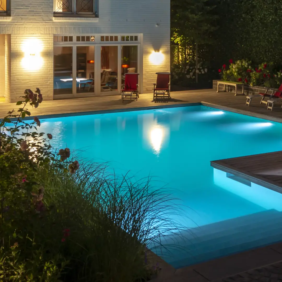 Blauer LivingPool mit ruhigem Wasser und reflektierenden Lichtern, umgeben von Terrassenfläche, Zierpflanzen und warmen Außenleuchten am Abend