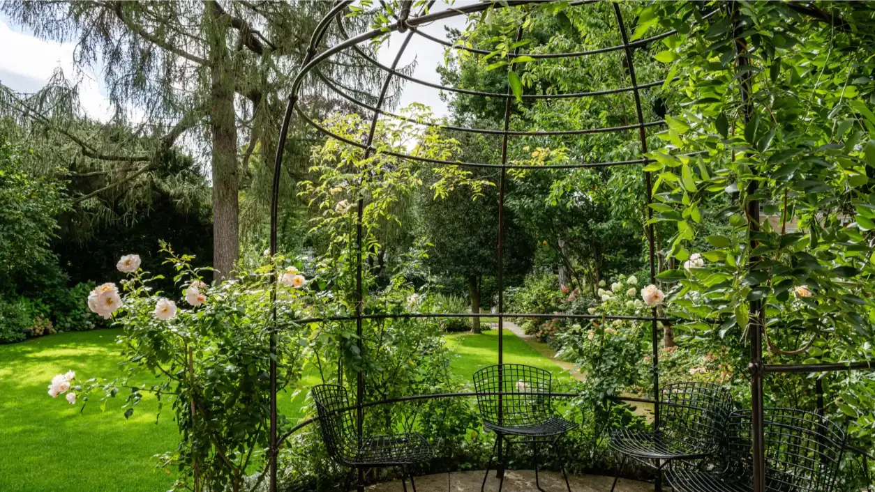 Unter einem filigranen Eisenpavillon mit Kletterrosen stehen mehrere schwarze Stühle in einem üppig grünen Garten mit Rasen, Bäumen und Blüten.