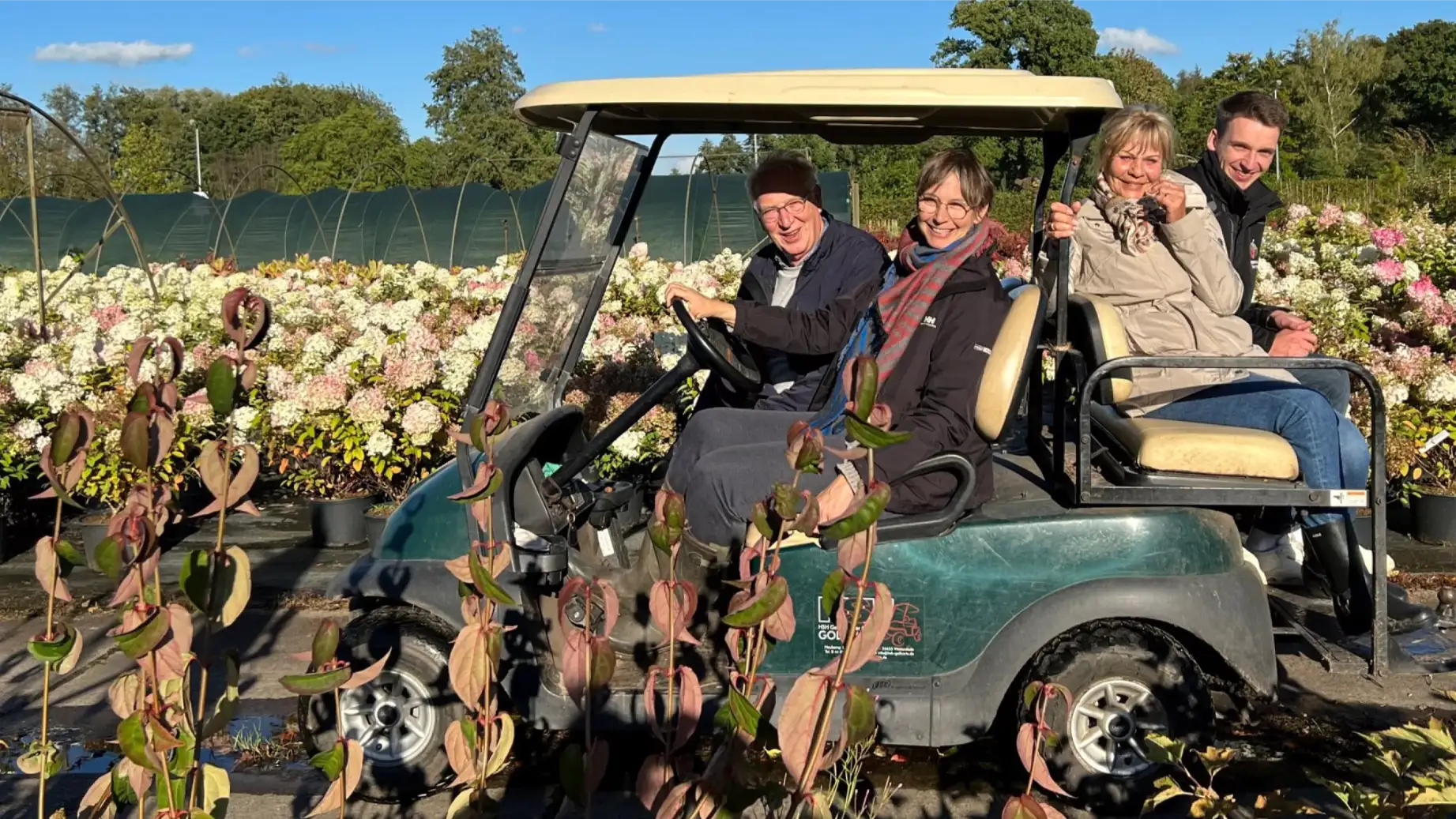 Vier Personen sitzen in einem Golfwagen, umgeben von blühenden Pflanzen in einem Garten.