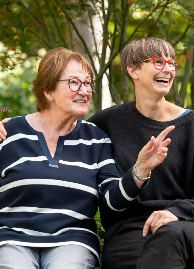 Zwei Frauen sitzen im grünen Garten und lachen gemeinsam, während die ältere Frau mit Brille und die jüngere Frau mit roten Brillen sprechen.