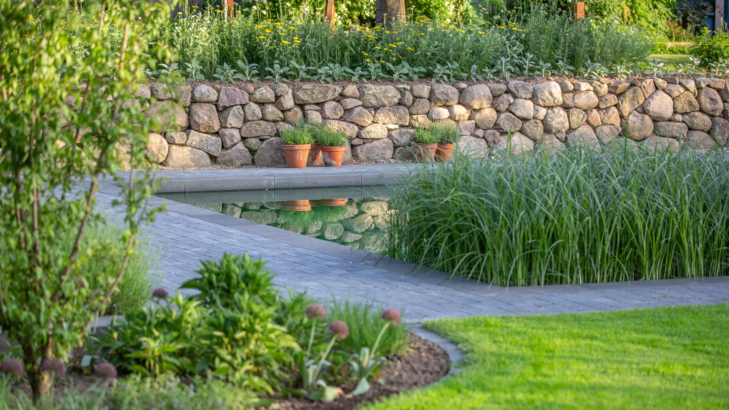 Garten mit Steinfassade, Wasserfläche und grünen Pflanzen in Töpfen.