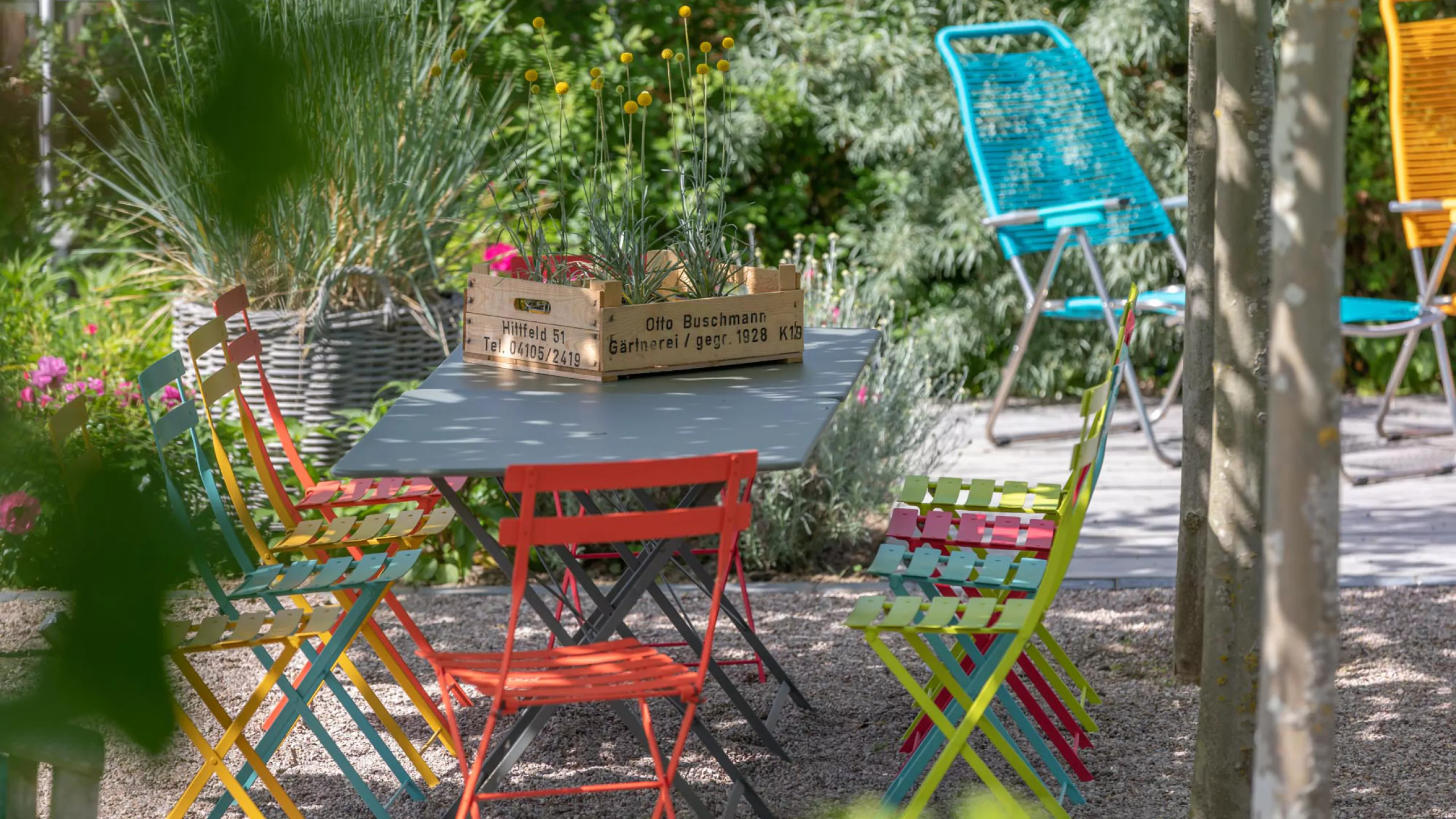 Bunte Gartenstühle stehen um einen grauen Bistrotisch zwischen üppigen Pflanzen, während im Hintergrund weitere farbige Sitzmöbel auf der Terrasse stehen.