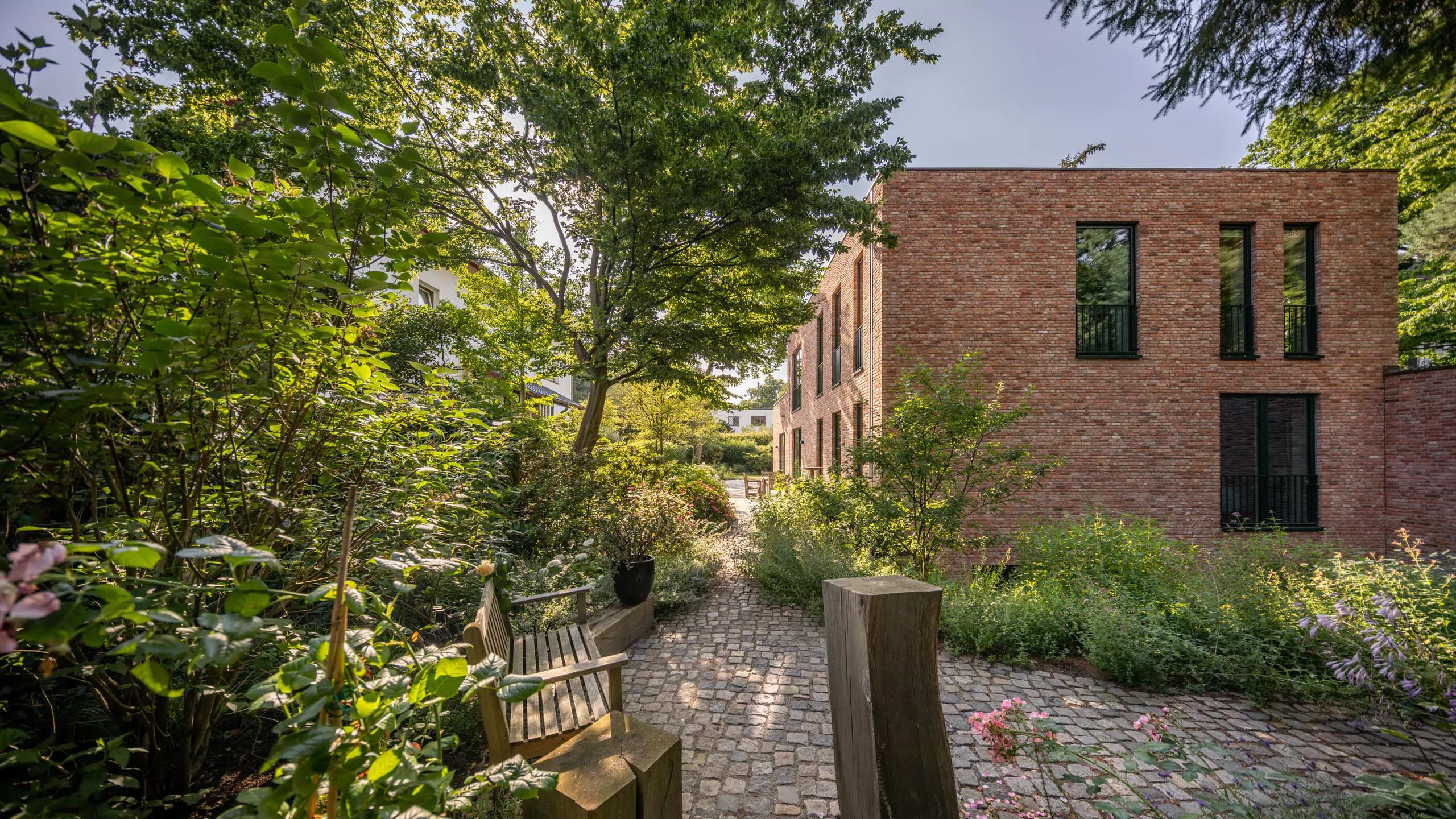 Gepflasterter Gartenweg am Backsteinhaus, Staudenbeete und Klinkerfassade Zwei moderne Backsteinhäuser umgeben von üppigem Grün und Bäumen in einem Garten.
