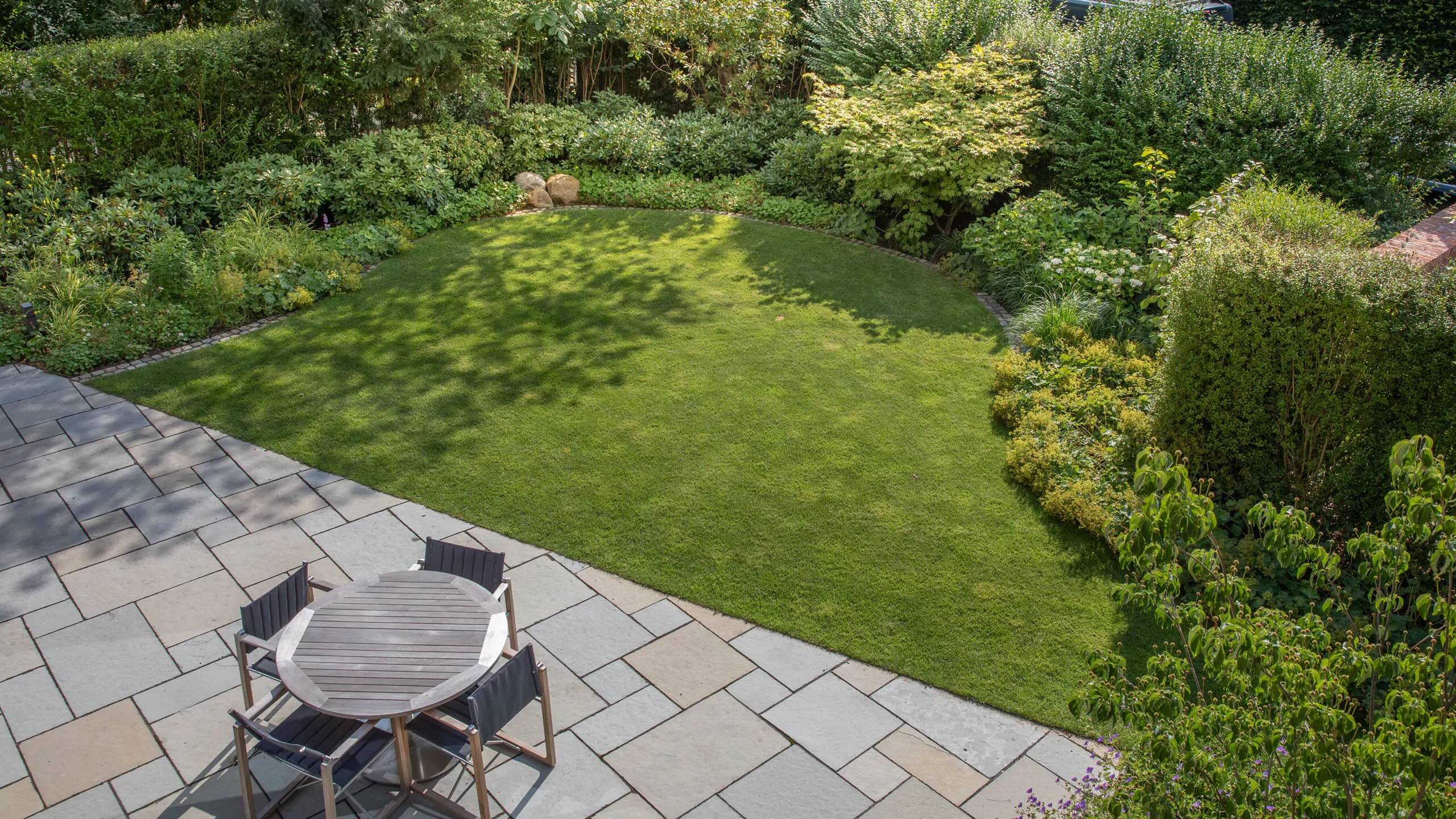 Terrassengestaltung mit Staudenbeet und gepflegter Rasenfläche im Garten Garten mit gepflegter Rasenfläche und Steinterrasse, umgeben von verschiedenen Pflanzen und Sträuchern.