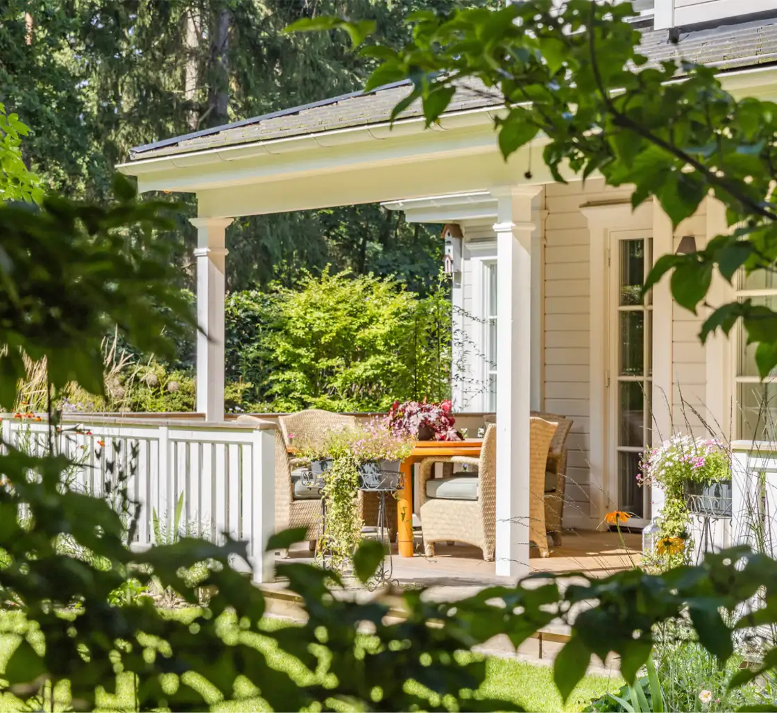 Beran Gärten Landhausveranda mit Korbstühlen und Blumen im Garten Gruppe von Mitarbeitern von Beran Gärten, die auf einer Wiese stehen, mit blauen Himmel und Wolken im Hintergrund.