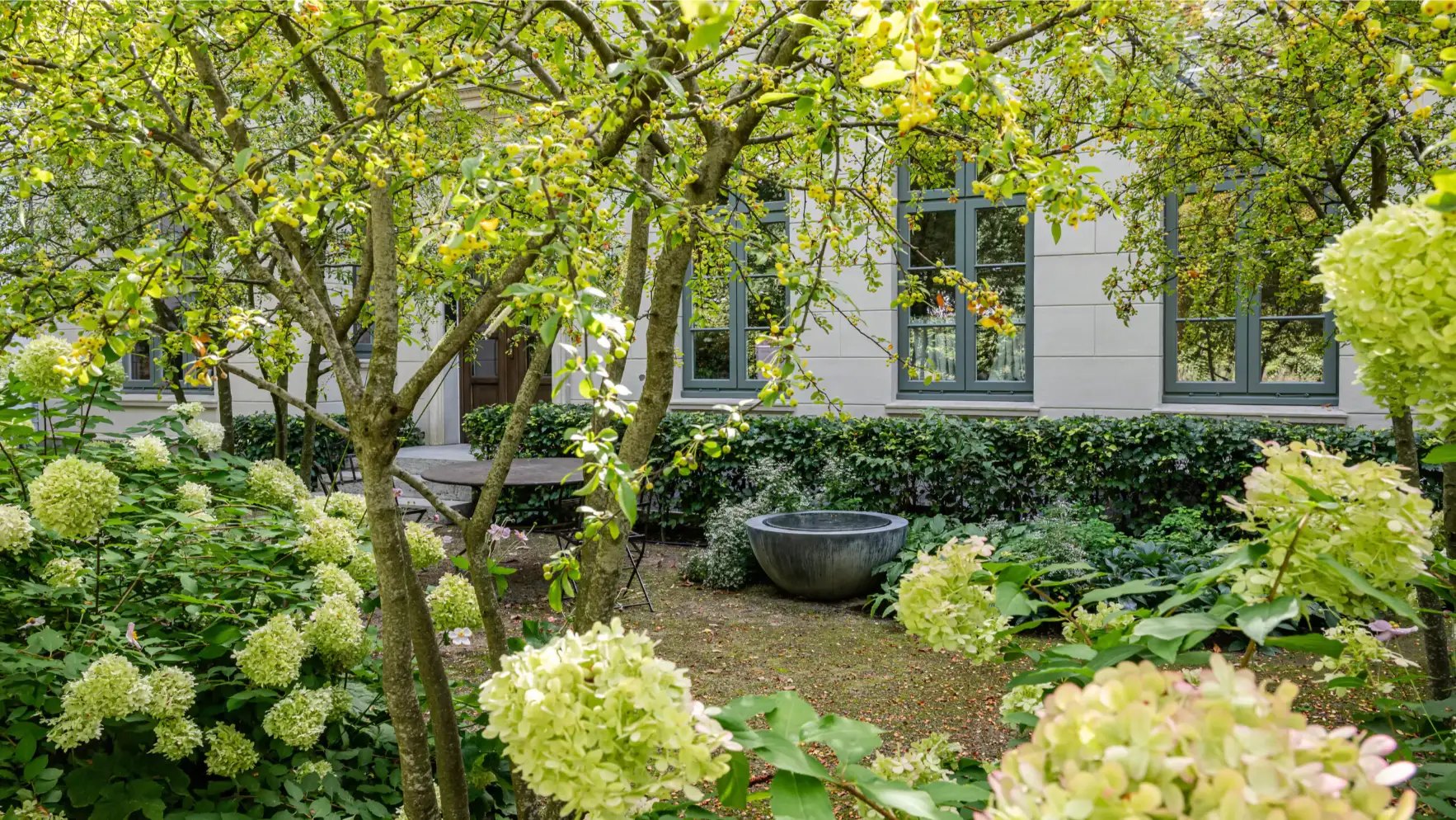 Zwischen Apfelbäumen und Hortensien steht ein rundes Steingefäß vor dem Haus von Beran Gärten, umgeben von dichtem, grünem Pflanzengrund und Hecken.