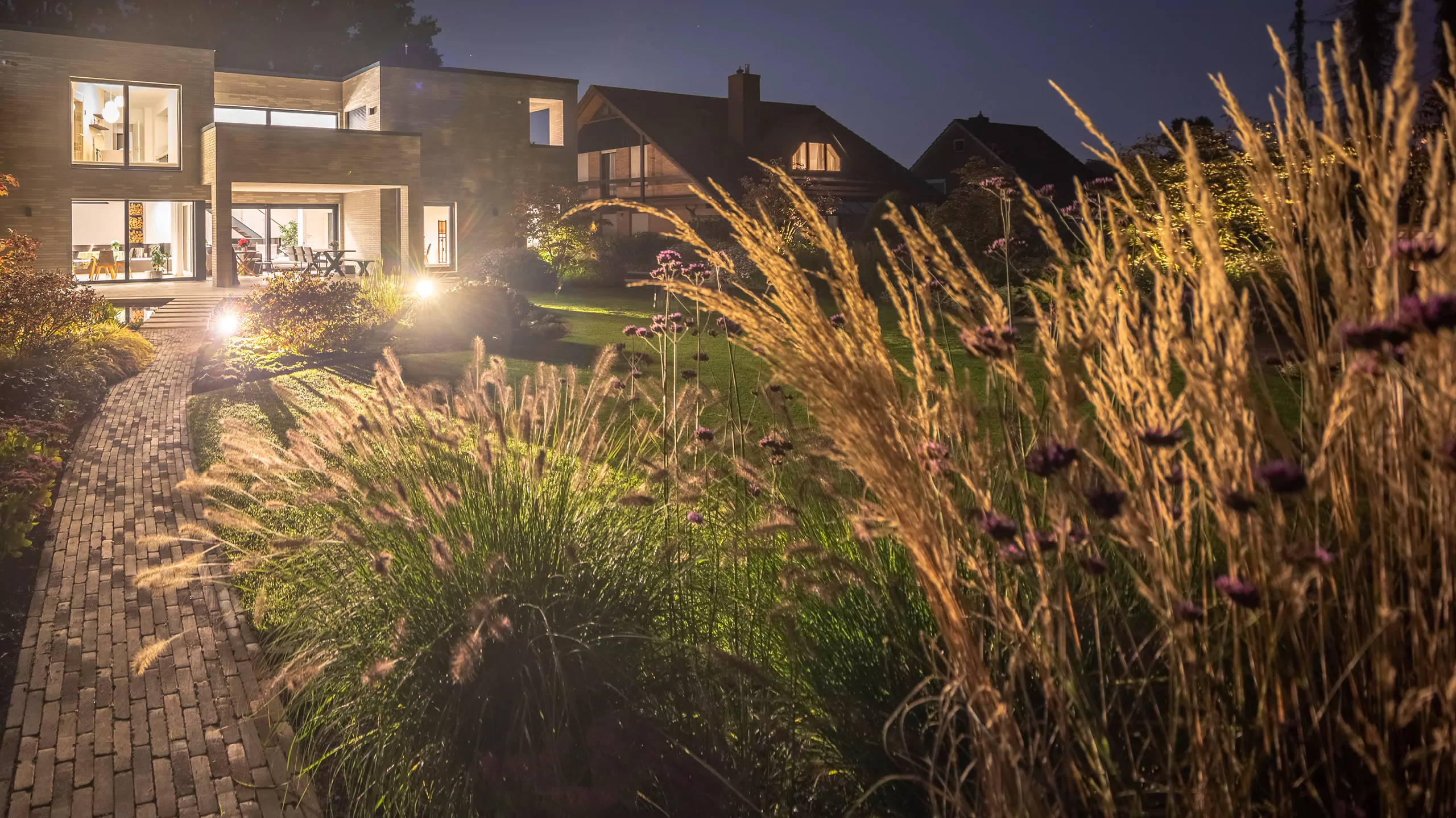 Ein moderner Garten von Beran Gärten leuchtet nachts mit Ziergräsern, Weg aus Pflastersteinen und warmen Lichtakzenten am Haus.