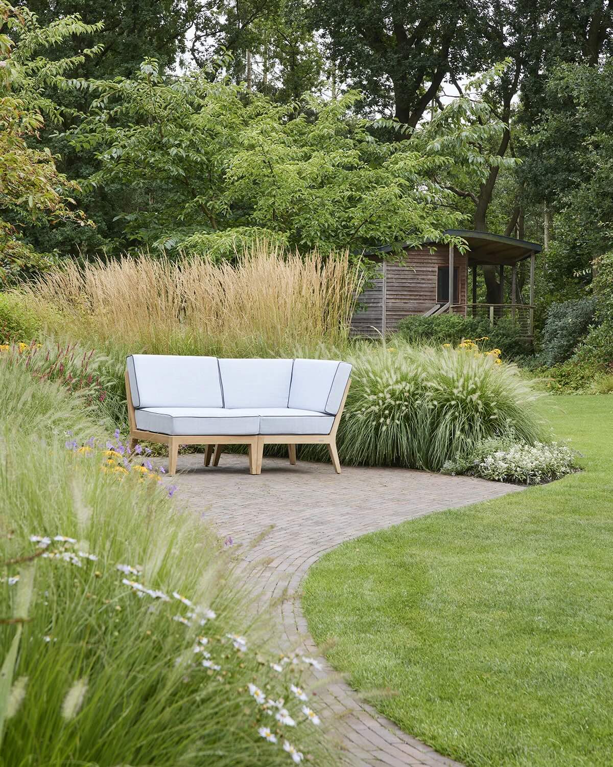 Gartengestaltung mit Holz-Baumhaus und hellem Loungemöbel bei Beran Gärten Ein helles Gartensofa steht auf einer Pflasterfläche vor üppigen Gräsern und einem kleinen Holzhaus in einem dicht bepflanzten Garten.